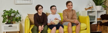 Üç çocuk, sarı bir kanepede oturup patlamış mısırlı film izliyor..