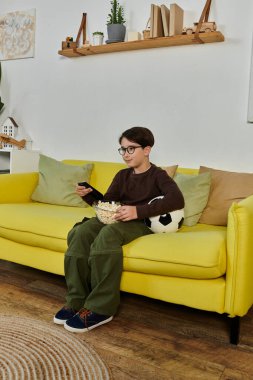 A boy watch TV on a yellow couch, sharing popcorn. clipart