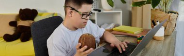 A young boy with Down syndrome uses a laptop at home, holding a coconut in his hand. clipart