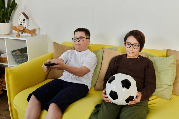 Два мальчика, один держит футбольный мяч, расслабиться на желтом диване в домашних условиях.