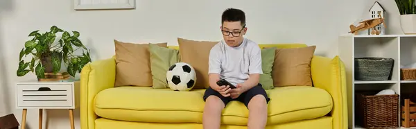 stock image A young boy sits on a yellow couch, holding a phone, with a soccer ball nearby.