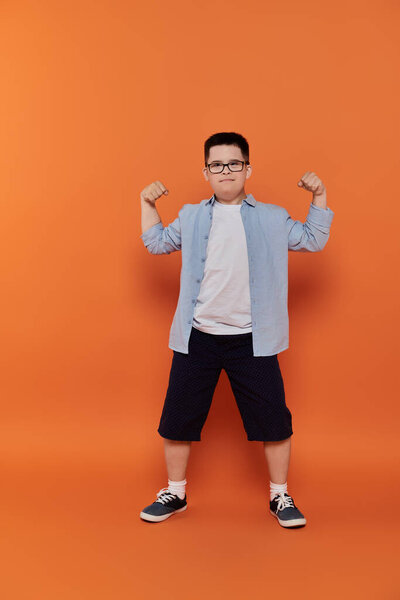 Мальчик с синдромом Дауна напрягает мышцы перед оранжевым фоном..