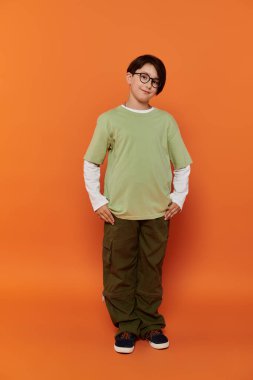 Genç bir çocuk, elleri kalçasında, yeşil gömlekli ve haki pantolonlu, kendinden emin bir şekilde ayakta duruyor..