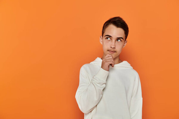 A young boy in a white hoodie gazes thoughtfully upwards against an orange background.