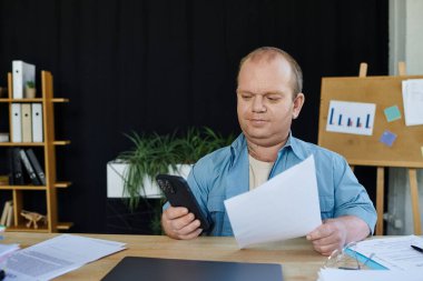 Kapsamlı bir adam masasında oturuyor, belgeleri gözden geçiriyor ve telefonuna bakıyor..