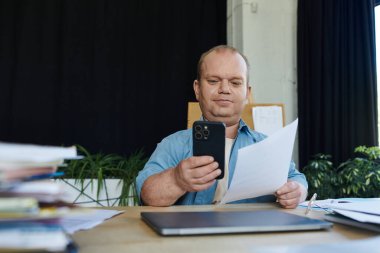 Kapsamlı bir adam masada oturuyor, elinde akıllı telefon tutarken belgeleri inceliyor..