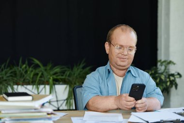 Kapsayıcı gözlüklü bir adam masada otururken akıllı telefonuna bakar..