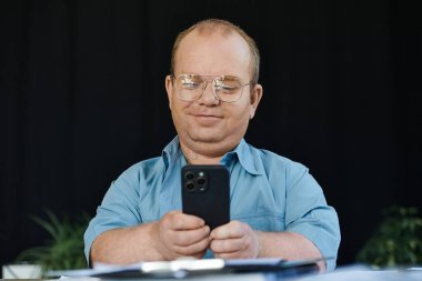 A man with inclusivity wearing glasses uses his smartphone in a dimly lit room. clipart