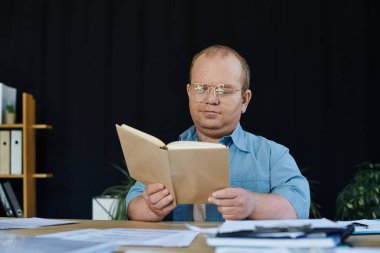 Kapsayıcı gözlüklü bir adam bir masada oturmuş, dikkatle kitap okuyor..