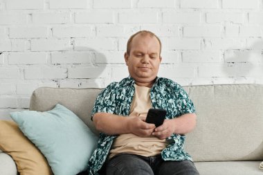 Kapsamlı bir adam beyaz tuğlalı arka planı olan bir kanepede oturur ve akıllı telefonuna bakar..
