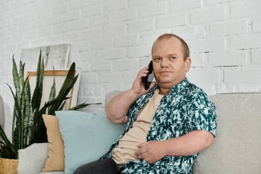 Çiçekli gömlek giyen bir adam kanepeye oturur ve bir telefon görüşmesinde konuşur..