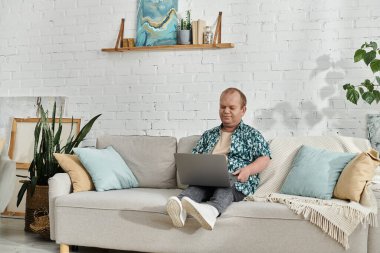 A man with inclusivity sits on a couch, working on a laptop in a bright living room. clipart