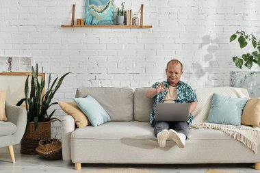 Kapsayıcı bir adam ev ofisinin kanepesinde oturmuş özenle dizüstü bilgisayarı üzerinde çalışıyor..