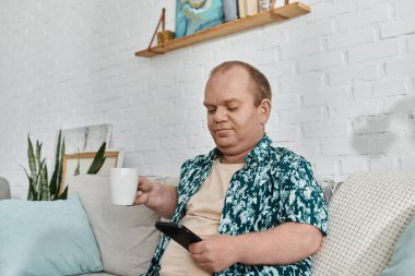 Kapsayıcı bir adam oturma odasında bir kanepeye oturur, bir fincan kahvenin tadını çıkarır ve telefonunu karıştırır..