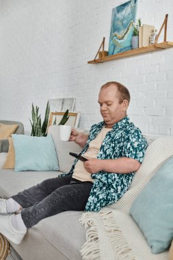 Çiçekli gömleği olan bir adam elinde telefon ve bir fincan kahveyle kanepede oturur..