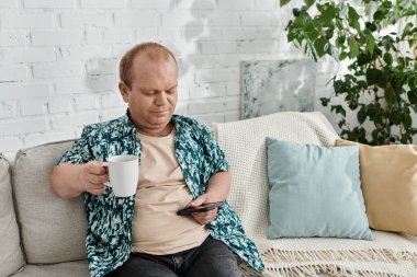 Kapsayıcı bir adam kanepeye oturur, elinde bir fincan kahve ve bir telefon ile..