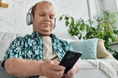 Kapsayıcı kulaklık takan bir adam bir kanepeye oturur, akıllı bir telefon tutar ve müzik dinler..