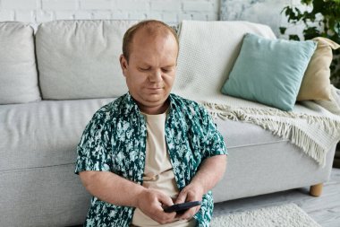 Şablonlu gömleği olan bir adam kanepenin önünde akıllı telefonunu kullanarak yere oturur..