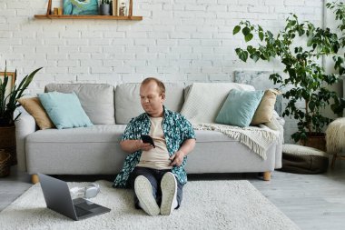 A man with inclusivity sits on a rug in a modern living room, using his phone. clipart