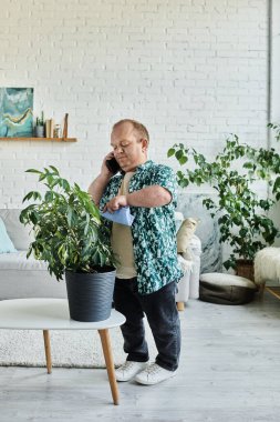 Kapsayıcılığı olan bir adam ev bitkisiyle ilgilenirken telefonuyla konuşuyor..