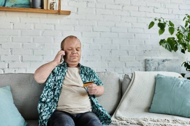 Kapsamlı bir adam kanepeye oturur, telefonuyla konuşur ve kredi kartı tutar..