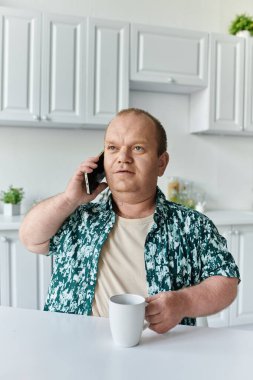 Yeşil desenli tişörtlü bir adam elinde beyaz bardakla telefonuyla konuşuyor..