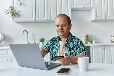 A man with inclusivity is sitting at a table in a kitchen, using a laptop and credit card to make a purchase online. clipart