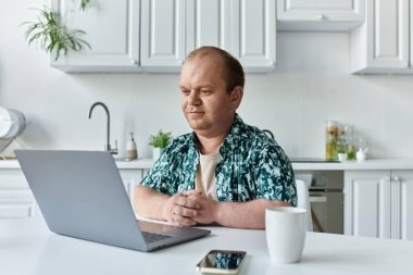 Kapsayıcılığı olan bir adam modern bir mutfaktaki bir masada oturup dizüstü bilgisayarla çalışır..