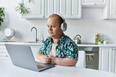 Kapsayıcı bir adam mutfaktaki bir masaya oturur, kulaklık takar ve dizüstü bilgisayarına odaklanır..