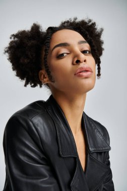 Siyah deri ceketli şık bir Afro-Amerikalı kadın gri arka planda poz veriyor..