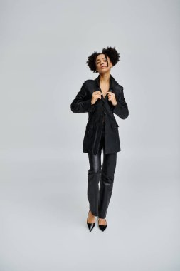 Siyah ceketli ve pantolonlu şık bir Afrikalı genç Amerikalı kadın gri bir fona karşı poz veriyor..