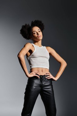 Beyaz askılı bluz giyen şık bir Afro-Amerikalı kadın gri bir fona karşı poz veriyor..