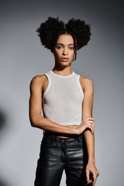 Beyaz atletli ve siyah deri pantolonlu genç bir Afro-Amerikalı kadın gri arka planda poz veriyor..