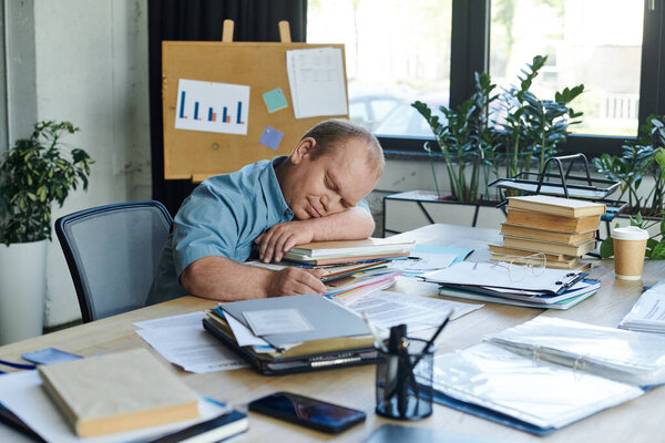 Человек с инклюзивностью кладет голову на стопку файлов, быстро дремлет за своим столом.