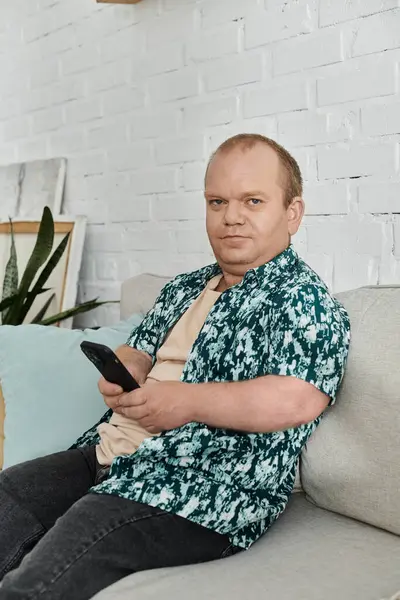 stock image A man with inclusivity sits on a couch, holding a phone and looking intently at the screen.