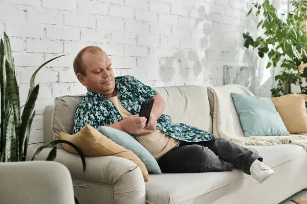 Stock image A man with inclusivity is sitting on a white couch in a bright, well-lit room, casually browsing his phone.