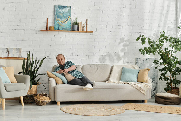 Человек с инклюзивностью сидит на сером диване в современном доме, расслабляется и пользуется телефоном.