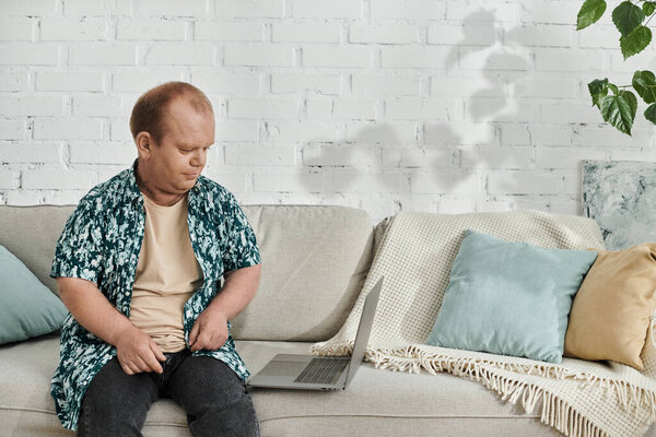 Человек с инклюзивностью сидит на диване, регулируя рубашку, с ноутбуком на диване рядом с ним.