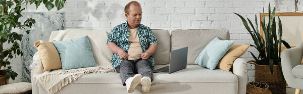 Мужчина с включённостью в узорчатой рубашке сидит на диване с ноутбуком и смотрит на экран.