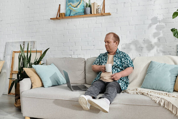 Человек с инклюзивностью сидит на диване, держа чашку кофе и глядя на ноутбук.