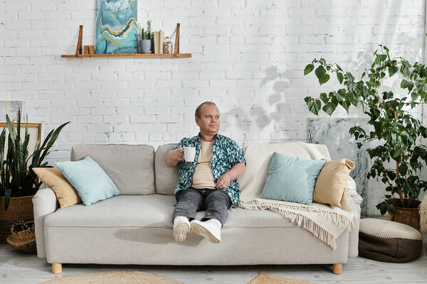 A man with inclusivity sits on a couch in a living room, enjoying a cup of coffee.