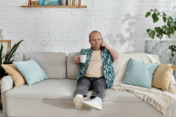 Человек с инклюзивностью сидит на диване, держа чашку кофе и разговаривая по телефону.
