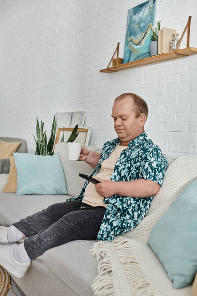 Мужчина с включённостью в цветочной рубашке сидит на диване, держа телефон и чашку кофе.