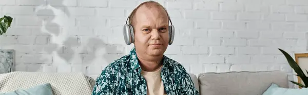 stock image A man with inclusivity sits on a couch with headphones on, listening to music in front of a white brick wall.