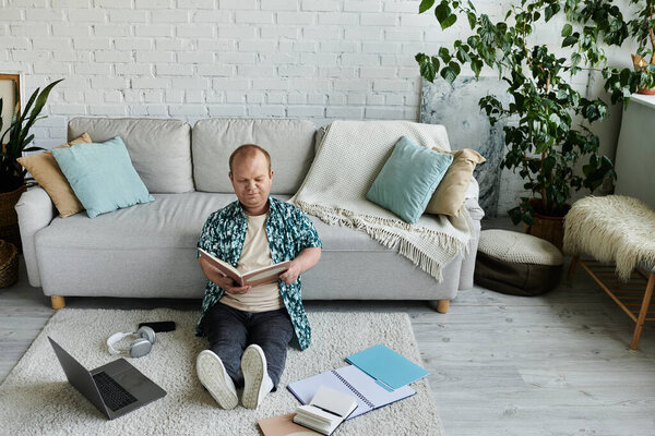 Человек с инклюзивностью сидит на ковре, читая книгу в гостиной.