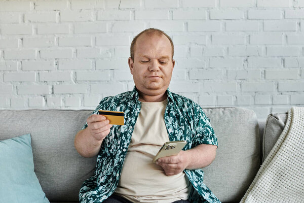 A man with inclusivity sits on a couch, holding a credit card and smartphone, likely making an online purchase.