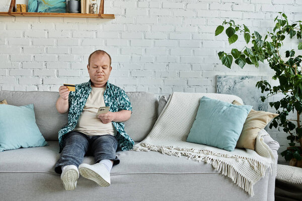 Человек с инклюзивностью сидит на диване, держа кредитку и используя смартфон.