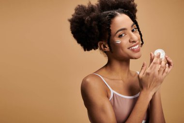Genç bir Afrikalı Amerikalı kadın, farklı ten tonlarının güzelliğini vurgulayarak, yüz nemlendiricisini uygularken kendine güvenerek gülümsüyor..
