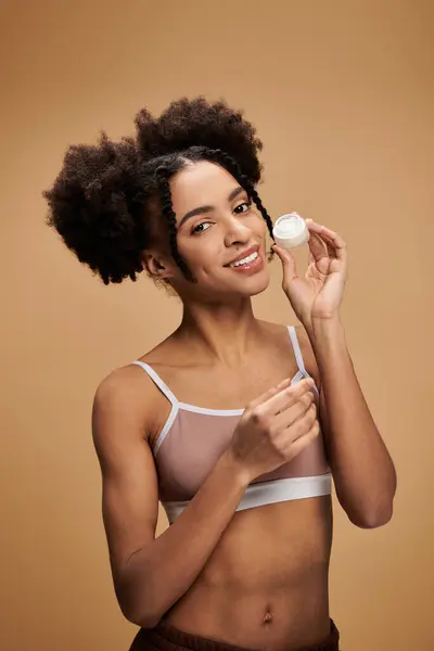 Stock image A young African American woman smiles brightly while holding a cream jar against a beige backdrop.