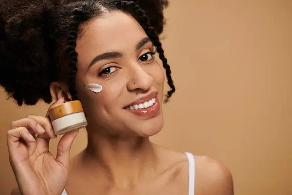 Stock image A young African American woman applies face cream to her cheek, smiling brightly. She is wearing a white tank top.